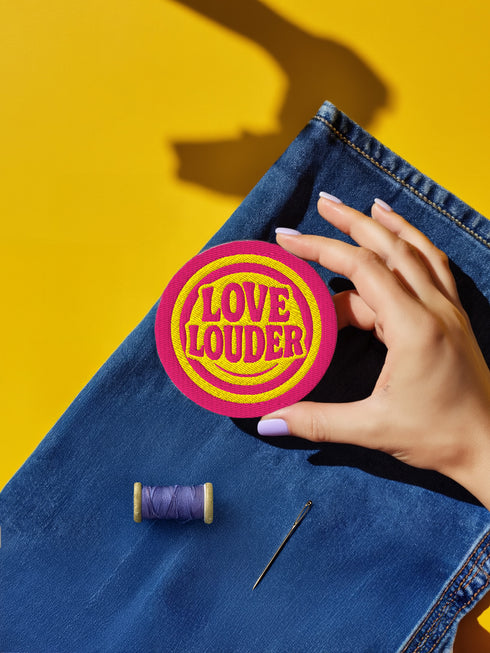 A round, 3-inch embroidered Love Louder patch designed with a vibrant, retro 70s aesthetic. The design features groovy, puffy-style text in a bright, sunny yellow thread, set against a bold magenta pink twill fabric background. The words 'LOVE LOUDER' are stacked to fill the center, encircled by concentric yellow rings that create a hypnotic, radiating effect. The entire circular patch is finished with a clean, durable merrowed border.