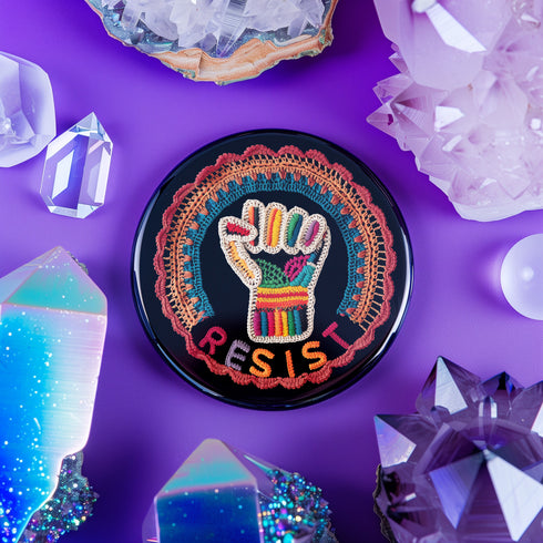 Round pinback metal button with an embroidered-style design on a black background. The center features a raised fist symbol stitched in a multicolored pattern, incorporating rainbow hues and folk-inspired textures. The fist is encircled by a vibrant, lace-like border in shades of blue, orange, and red, resembling intricate embroidery. Below the fist, the word "RESIST" is spelled out in bold, stitched-textured letters in red and orange. The button has a silver metal back with a secure pin closure.