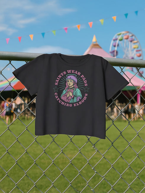 Black cropped t-shirt featuring a colorful, stylized saint figure with pink hair, wearing a helmet and pads. Surrounded by radiant lines and the phrase “Saints Wear Pads Catching Elbows” in bold pastel text. The design has a vintage punk-meets-holy aesthetic with star accents and a retro vibe. Ideal for roller skate girls, derby athletes, and goth skater fans. Available in sizes XS–2XL with raw hem details.