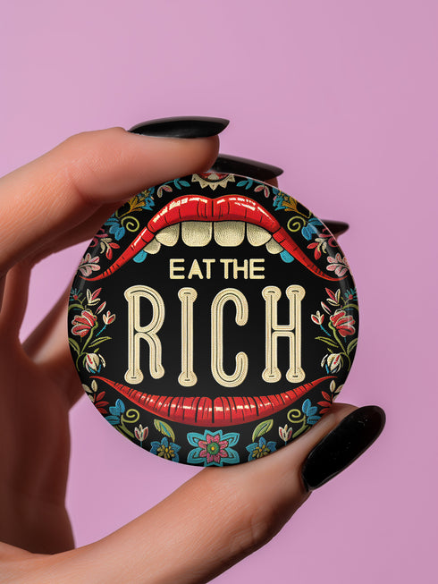 A round pin-back button with a bold, embroidered-style design featuring the phrase "EAT THE RICH" in large, vintage-style lettering. The typography is framed by a set of open red lips with detailed stitching, revealing white teeth. Surrounding the mouth are intricate floral patterns in shades of blue, red, and green, adding a decorative, folk-art-inspired touch. The button has a black background, making the details and lettering stand out