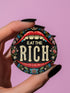 A round pin-back button with a bold, embroidered-style design featuring the phrase "EAT THE RICH" in large, vintage-style lettering. The typography is framed by a set of open red lips with detailed stitching, revealing white teeth. Surrounding the mouth are intricate floral patterns in shades of blue, red, and green, adding a decorative, folk-art-inspired touch. The button has a black background, making the details and lettering stand out
