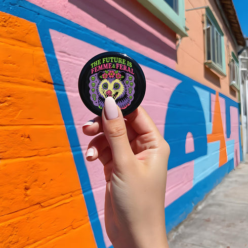 A round, black pinback button with a high-gloss finish features the text "THE FUTURE IS FEMME & FERAL" in a retro font of lime green and pastel pink. In the center, a screaming possum with wild eyes and a pink-lined mouth is illustrated in a pastel goth style with purple, yellow, and pink fur. The possum wears a pink flower crown. The illustration is framed by a circle of small skulls and pink Venus (female) symbols on a purple background. This is a piece of feminist possumcore flair.