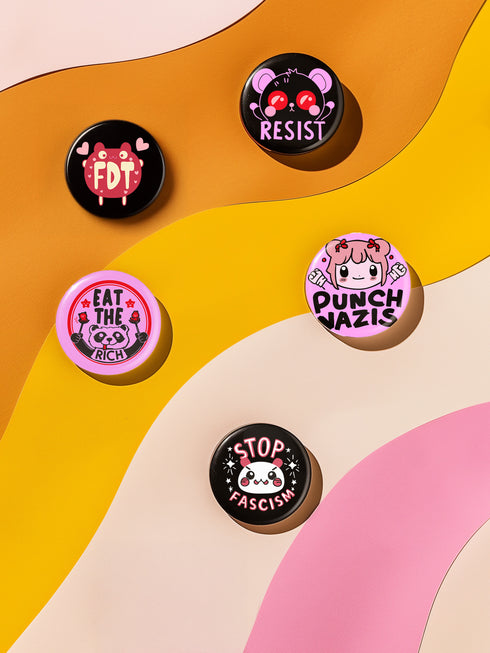 A set of five glossy round pinback buttons with pastel goth and kawaii punk designs, featuring slogans like "Eat The Rich" and "Stop Fascism" in bold pink and black. Each pin has a cute character with a rebellious message.