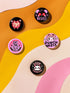 A set of five glossy round pinback buttons with pastel goth and kawaii punk designs, featuring slogans like "Eat The Rich" and "Stop Fascism" in bold pink and black. Each pin has a cute character with a rebellious message.