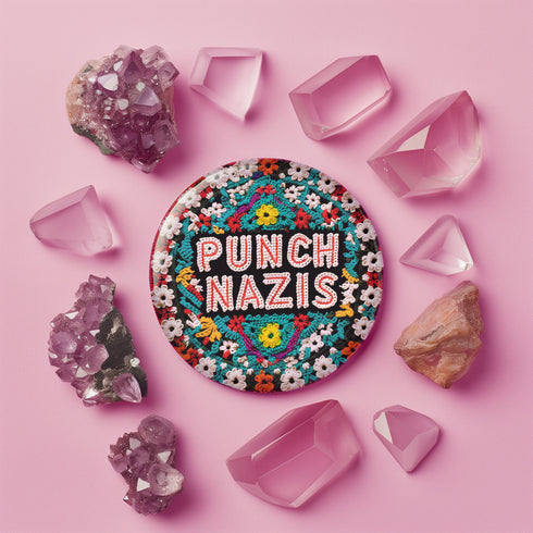 Round pinback metal button with an embroidered-style design on a black background. The center displays the phrase "PUNCH NAZIS" in bold, white stitched-textured lettering with red accents. Surrounding the text is an elaborate folk-inspired floral pattern, densely packed with embroidered-style flowers in shades of red, yellow, blue, pink, and white. The intricate design includes symmetrical lace-like stitching and decorative motifs, creating a rich, tapestry-like effect.