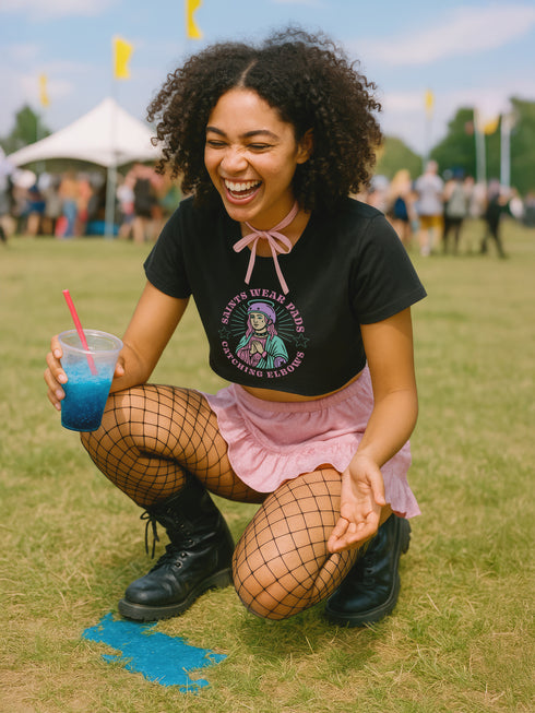 Black cropped t-shirt featuring a colorful, stylized saint figure with pink hair, wearing a helmet and pads. Surrounded by radiant lines and the phrase “Saints Wear Pads Catching Elbows” in bold pastel text. The design has a vintage punk-meets-holy aesthetic with star accents and a retro vibe. Ideal for roller skate girls, derby athletes, and goth skater fans. Available in sizes XS–2XL with raw hem details.