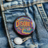 Round black pinback button with neon embroidery-style waves surrounding bold yellow and red "DISOBEY" text.