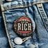A round pin-back button with a bold, embroidered-style design featuring the phrase "EAT THE RICH" in large, vintage-style lettering. The typography is framed by a set of open red lips with detailed stitching, revealing white teeth. Surrounding the mouth are intricate floral patterns in shades of blue, red, and green, adding a decorative, folk-art-inspired touch. The button has a black background, making the details and lettering stand out