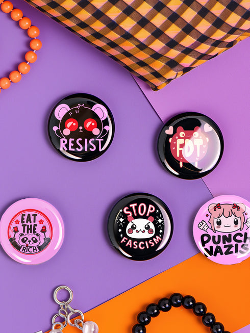 A set of five glossy round pinback buttons with pastel goth and kawaii punk designs, featuring slogans like "Eat The Rich" and "Stop Fascism" in bold pink and black. Each pin has a cute character with a rebellious message.