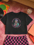 Black cropped t-shirt featuring a colorful, stylized saint figure with pink hair, wearing a helmet and pads. Surrounded by radiant lines and the phrase “Saints Wear Pads Catching Elbows” in bold pastel text. The design has a vintage punk-meets-holy aesthetic with star accents and a retro vibe. Ideal for roller skate girls, derby athletes, and goth skater fans. Available in sizes XS–2XL with raw hem details.