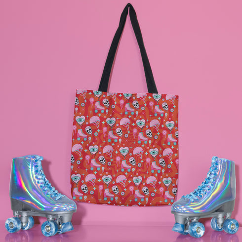 Tote bag with black handles featuring a vibrant red background covered in pastel pink and blue illustrations of skulls, roller skates, eyes, and geometric shapes. Displayed on an orange surface with dramatic shadows.