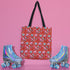 Tote bag with black handles featuring a vibrant red background covered in pastel pink and blue illustrations of skulls, roller skates, eyes, and geometric shapes. Displayed on an orange surface with dramatic shadows.