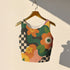 Cropped racerback tank top featuring bold retro floral print with orange and yellow mod flowers, green leaves, black and white checkerboard pattern, and abstract wavy designs. Fitted athletic style crop top in vibrant 70s inspired botanical print with performance fabric construction. Women's festival rave wear cropped camisole with psychedelic flower power design perfect for summer concerts, Y2K fashion looks, and alternative streetwear styling.