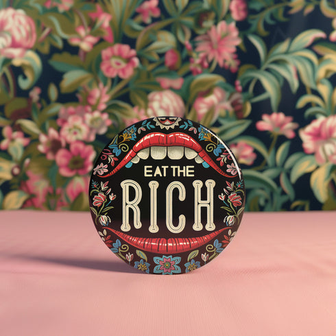 A round pin-back button with a bold, embroidered-style design featuring the phrase "EAT THE RICH" in large, vintage-style lettering. The typography is framed by a set of open red lips with detailed stitching, revealing white teeth. Surrounding the mouth are intricate floral patterns in shades of blue, red, and green, adding a decorative, folk-art-inspired touch. The button has a black background, making the details and lettering stand out