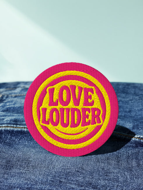 A round, 3-inch embroidered Love Louder patch designed with a vibrant, retro 70s aesthetic. The design features groovy, puffy-style text in a bright, sunny yellow thread, set against a bold magenta pink twill fabric background. The words 'LOVE LOUDER' are stacked to fill the center, encircled by concentric yellow rings that create a hypnotic, radiating effect. The entire circular patch is finished with a clean, durable merrowed border.