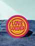 A round, 3-inch embroidered Love Louder patch designed with a vibrant, retro 70s aesthetic. The design features groovy, puffy-style text in a bright, sunny yellow thread, set against a bold magenta pink twill fabric background. The words 'LOVE LOUDER' are stacked to fill the center, encircled by concentric yellow rings that create a hypnotic, radiating effect. The entire circular patch is finished with a clean, durable merrowed border.