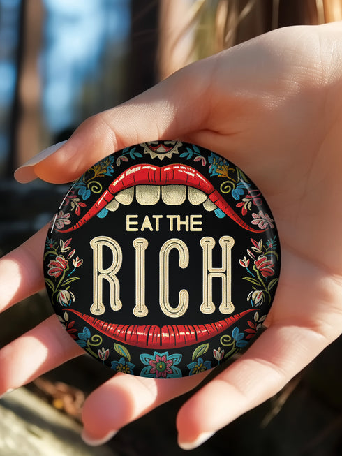 A round pin-back button with a bold, embroidered-style design featuring the phrase "EAT THE RICH" in large, vintage-style lettering. The typography is framed by a set of open red lips with detailed stitching, revealing white teeth. Surrounding the mouth are intricate floral patterns in shades of blue, red, and green, adding a decorative, folk-art-inspired touch. The button has a black background, making the details and lettering stand out