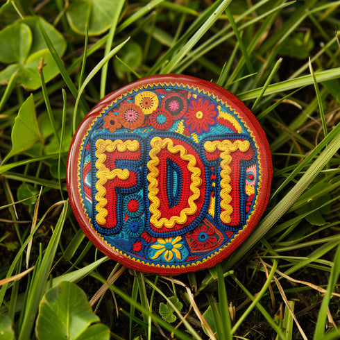 Round pinback metal button featuring an embroidered-style design on a vibrant, multicolored background. The bold, textured letters "FDT" are prominently displayed in red with intricate golden-yellow stitching details, outlined in dark blue. Surrounding the text is an ornate floral pattern with embroidered-style flowers in shades of red, orange, blue, and yellow, densely arranged for a rich, decorative effect. The outer edge is bordered with a zigzag stitching effect in red and blue. The button's back is sil