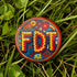 Round pinback metal button featuring an embroidered-style design on a vibrant, multicolored background. The bold, textured letters "FDT" are prominently displayed in red with intricate golden-yellow stitching details, outlined in dark blue. Surrounding the text is an ornate floral pattern with embroidered-style flowers in shades of red, orange, blue, and yellow, densely arranged for a rich, decorative effect. The outer edge is bordered with a zigzag stitching effect in red and blue. The button's back is sil
