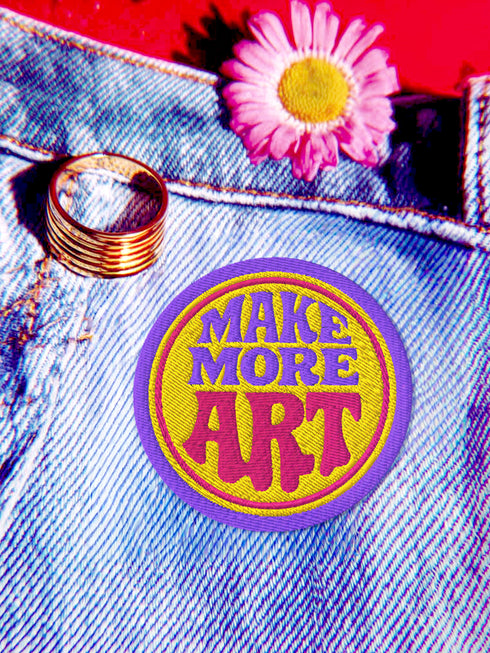 A round embroidered patch with the words "MAKE MORE ART" in a retro 70s psychedelic font. The word "ART" is magenta pink, "MAKE MORE" is purple, and they are set against a bright yellow circular background. The entire design is encircled by a thin pink line and a thick purple border. This 3-inch iron-on applique is a colorful, groovy accessory for DIY projects, perfect for personalizing jackets, backpacks, or denim vests. It serves as an inspirational artist gift with a vintage aesthetic.