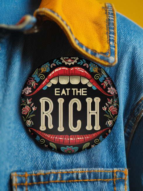 A round pin-back button with a bold, embroidered-style design featuring the phrase "EAT THE RICH" in large, vintage-style lettering. The typography is framed by a set of open red lips with detailed stitching, revealing white teeth. Surrounding the mouth are intricate floral patterns in shades of blue, red, and green, adding a decorative, folk-art-inspired touch. The button has a black background, making the details and lettering stand out