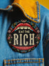 A round pin-back button with a bold, embroidered-style design featuring the phrase "EAT THE RICH" in large, vintage-style lettering. The typography is framed by a set of open red lips with detailed stitching, revealing white teeth. Surrounding the mouth are intricate floral patterns in shades of blue, red, and green, adding a decorative, folk-art-inspired touch. The button has a black background, making the details and lettering stand out