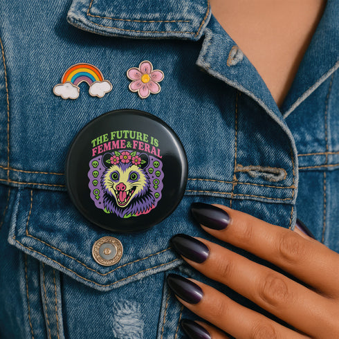 A round, black pinback button with a high-gloss finish features the text "THE FUTURE IS FEMME & FERAL" in a retro font of lime green and pastel pink. In the center, a screaming possum with wild eyes and a pink-lined mouth is illustrated in a pastel goth style with purple, yellow, and pink fur. The possum wears a pink flower crown. The illustration is framed by a circle of small skulls and pink Venus (female) symbols on a purple background. This is a piece of feminist possumcore flair.