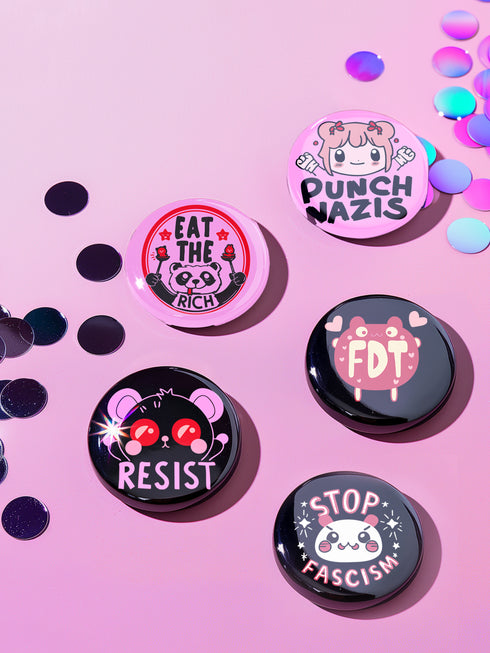 A set of five glossy round pinback buttons with pastel goth and kawaii punk designs, featuring slogans like "Eat The Rich" and "Stop Fascism" in bold pink and black. Each pin has a cute character with a rebellious message.