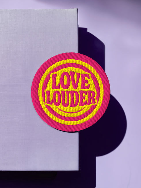 A round, 3-inch embroidered Love Louder patch designed with a vibrant, retro 70s aesthetic. The design features groovy, puffy-style text in a bright, sunny yellow thread, set against a bold magenta pink twill fabric background. The words 'LOVE LOUDER' are stacked to fill the center, encircled by concentric yellow rings that create a hypnotic, radiating effect. The entire circular patch is finished with a clean, durable merrowed border.
