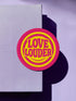 A round, 3-inch embroidered Love Louder patch designed with a vibrant, retro 70s aesthetic. The design features groovy, puffy-style text in a bright, sunny yellow thread, set against a bold magenta pink twill fabric background. The words 'LOVE LOUDER' are stacked to fill the center, encircled by concentric yellow rings that create a hypnotic, radiating effect. The entire circular patch is finished with a clean, durable merrowed border.