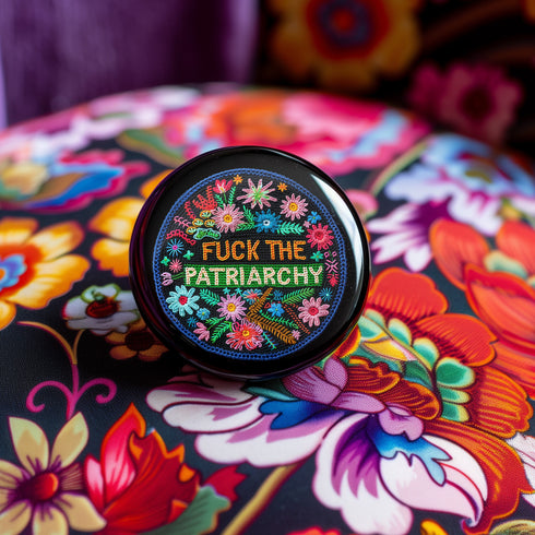 Circular black pinback button with "FUCK THE PATRIARCHY" in orange and mint green text, surrounded by embroidery-style flowers in pink, blue, and red with green foliage and blue border. Folk art feminist protest pin with glossy finish and safety pin backing in 2" and 3" sizes.