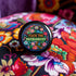 Circular black pinback button with "FUCK THE PATRIARCHY" in orange and mint green text, surrounded by embroidery-style flowers in pink, blue, and red with green foliage and blue border. Folk art feminist protest pin with glossy finish and safety pin backing in 2" and 3" sizes.