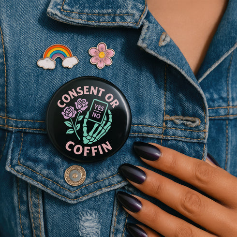 A round black enamel pinback button reading “Consent Or Coffin” in pastel-pink text above and below a mint-green skeleton hand holding a tiny phone interface labeled “YES/NO,” flanked by two stylized pink roses, on a textured purple background.