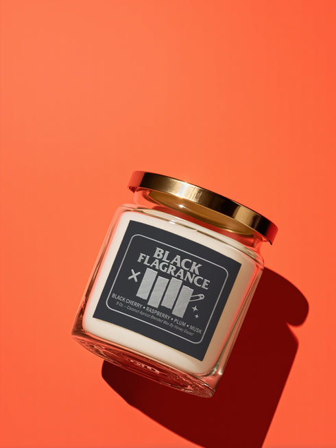 A 9-ounce hand-poured candle in a clear, cylindrical glass jar with a slight taper at the base. The wax is a creamy, solid off-white color, filling the jar to just below the rim where a single white cotton wick is centered. The label features the words "BLACK FLAGRANCE" in a bold, white, sans-serif font. Below this, a stylized graphic mimics a famous punk band logo with four vertical white bars. The candle is topped with a shiny, metallic gold-colored lid.