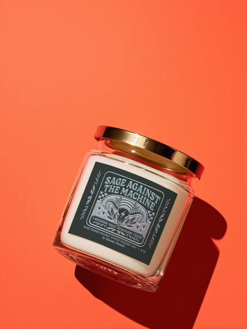 A 9-ounce, hand-poured scented candle in a transparent glass vessel. The candle contains a cotton wick and is filled with a creamy, off-white coconut apricot blended wax. It features a dark, matte finish label with a silver metallic emblem of a ram skull and stylized foliage, with the text "Sage Against The Machine" centered above the scent notes. A shiny gold lid sits atop the glass jar.