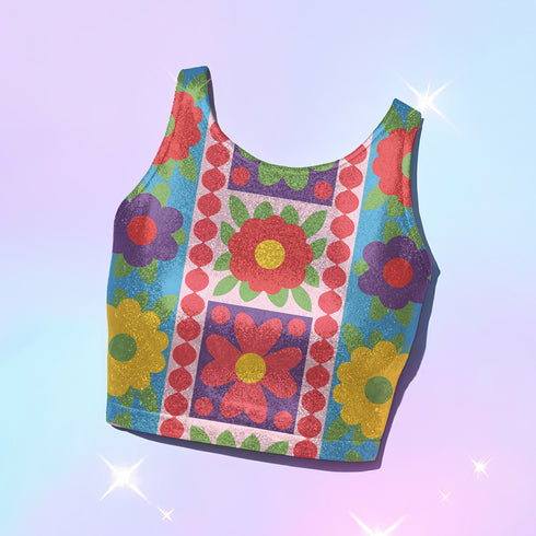 A high-resolution product photo of a body-hugging, 2D printed crop top with a 1970s-inspired floral patchwork pattern. The design consists of vibrant squares in cobalt blue, pale lavender, and deep violet, separated by pink borders with red dots. Central motifs include yellow daisies with orange centers and green-magenta floral mandalas. The top is made of a smooth, high-stretch performance knit fabric with curved back armholes and a cropped waistline.