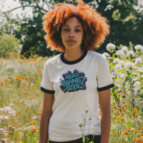 Off-white ringer t-shirt with navy contrast trim on the collar and sleeve cuffs. The front features a bold graffiti-style graphic with the phrase "READ BANNED BOOKS" in vibrant turquoise, layered over a stack of illustrated books in dark navy and black. The hand-drawn lettering and ink-splatter details give the design a rebellious, street-art-inspired aesthetic. The t-shirt has a vintage-inspired fit, made from soft cotton fabric with a slightly relaxed silhouette. The navy trim complements the graphic, add