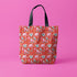 Tote bag with black handles featuring a vibrant red background covered in pastel pink and blue illustrations of skulls, roller skates, eyes, and geometric shapes. Displayed on an orange surface with dramatic shadows.