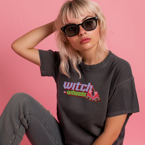 Gray t-shirt with "Witch on Wheels" text in purple and green, featuring a red roller skate graphic. The vintage-style design has a slightly faded look on a relaxed-fit unisex tee.