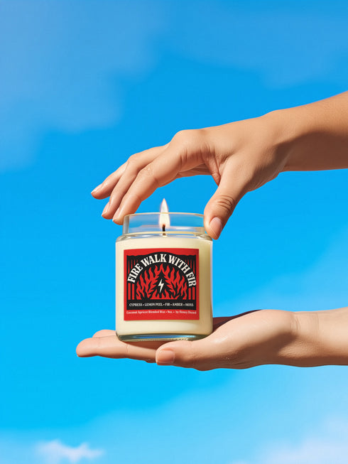 A 9-ounce hand-poured candle inspired by the TV show Twin Peaks, named "Fire Walk With Fir." The candle is made from a creamy, off-white coconut apricot blended wax, housed in a clear glass vessel with a classic, slightly tapered shape. A prominent matte-finish label on the front features the title in a stylized font over a red and black graphic with flame and fir tree motifs. The label also lists scent notes of cypress, lemon peel, fir, amber, and moss.