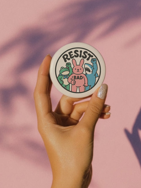 A round white pinback button with the word 'RESIST' in a bold, black, arched font. Below the text stand three cute cartoon characters raising their fists in protest: a green frog with a blue bandana, a pink bunny with '/BAD' on its chest, and a blue shark with sharp teeth. The activist pin has a glossy, protective mylar finish and a durable metal shell, designed as political statement flair for jackets, bags, and backpacks. This punk-inspired, kawaii- badge is a symbol of joyful rebellion.