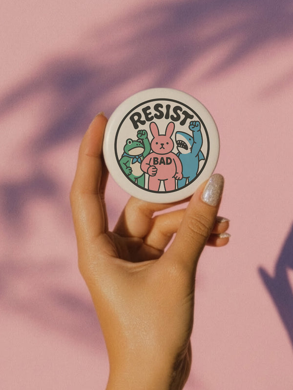 A round white pinback button with the word 'RESIST' in a bold, black, arched font. Below the text stand three cute cartoon characters raising their fists in protest: a green frog with a blue bandana, a pink bunny with '/BAD' on its chest, and a blue shark with sharp teeth. The activist pin has a glossy, protective mylar finish and a durable metal shell, designed as political statement flair for jackets, bags, and backpacks. This punk-inspired, kawaii- badge is a symbol of joyful rebellion.