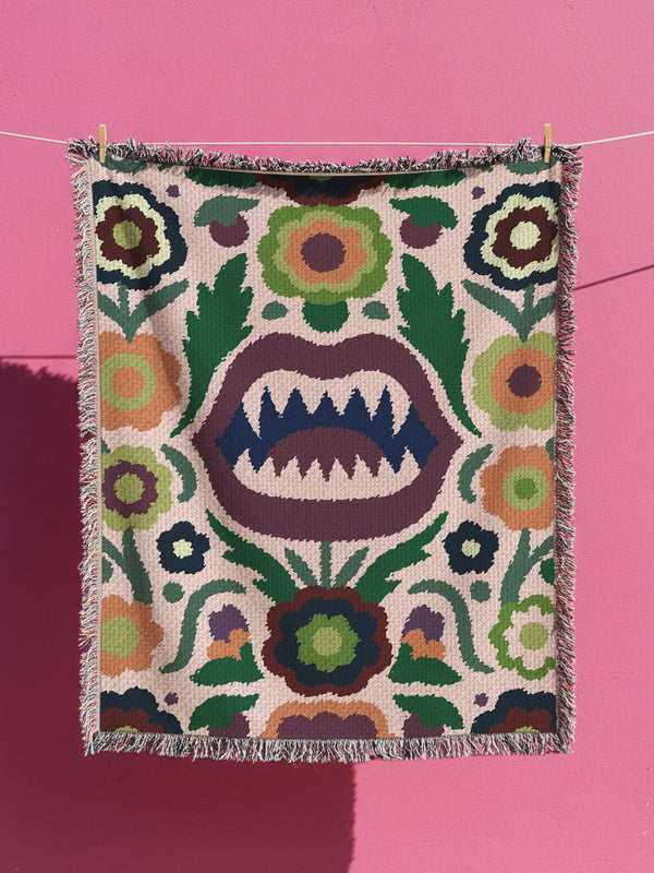 100% cotton woven throw blanket featuring a bold folk-art monster mouth with sharp fangs framed by retro floral and leaf motifs in green, plum, navy, gold, and sage on a warm neutral ground. Finished with multicolor fringe on all sides. Cozy, medium-weight jacquard weave. Available in three sizes for sofa, bed, or wall-hanging; perfect for gothic boho, alt, and maximalist home décor.
