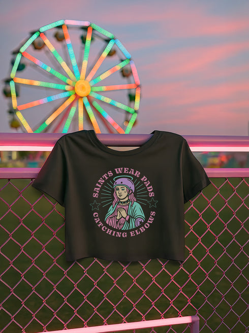 Black cropped t-shirt featuring a colorful, stylized saint figure with pink hair, wearing a helmet and pads. Surrounded by radiant lines and the phrase “Saints Wear Pads Catching Elbows” in bold pastel text. The design has a vintage punk-meets-holy aesthetic with star accents and a retro vibe. Ideal for roller skate girls, derby athletes, and goth skater fans. Available in sizes XS–2XL with raw hem details.