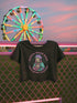 Black cropped t-shirt featuring a colorful, stylized saint figure with pink hair, wearing a helmet and pads. Surrounded by radiant lines and the phrase “Saints Wear Pads Catching Elbows” in bold pastel text. The design has a vintage punk-meets-holy aesthetic with star accents and a retro vibe. Ideal for roller skate girls, derby athletes, and goth skater fans. Available in sizes XS–2XL with raw hem details.