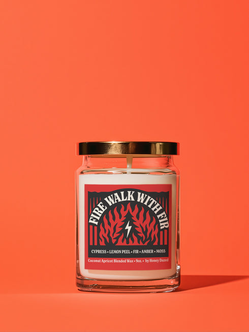 A 9-ounce hand-poured candle inspired by the TV show Twin Peaks, named "Fire Walk With Fir." The candle is made from a creamy, off-white coconut apricot blended wax, housed in a clear glass vessel with a classic, slightly tapered shape. A prominent matte-finish label on the front features the title in a stylized font over a red and black graphic with flame and fir tree motifs. The label also lists scent notes of cypress, lemon peel, fir, amber, and moss.
