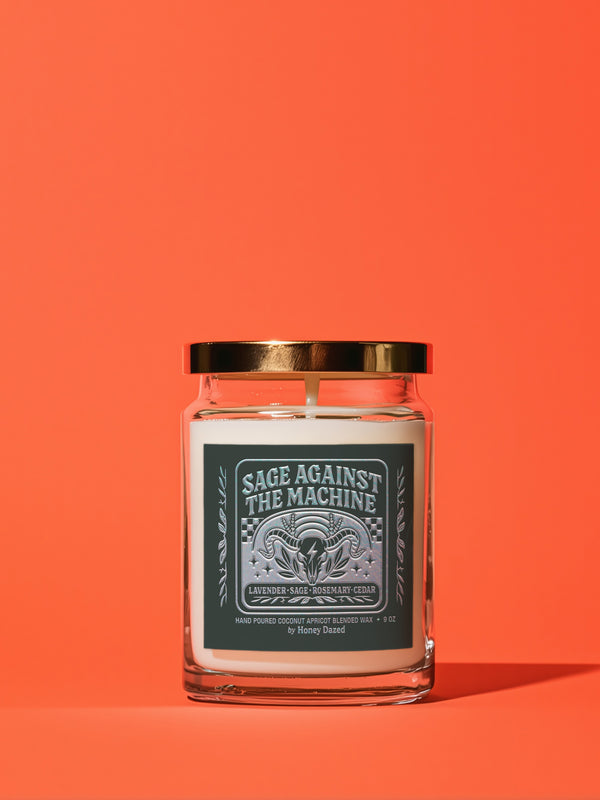 A 9-ounce, hand-poured scented candle in a transparent glass vessel. The candle contains a cotton wick and is filled with a creamy, off-white coconut apricot blended wax. It features a dark, matte finish label with a silver metallic emblem of a ram skull and stylized foliage, with the text "Sage Against The Machine" centered above the scent notes. A shiny gold lid sits atop the glass jar.