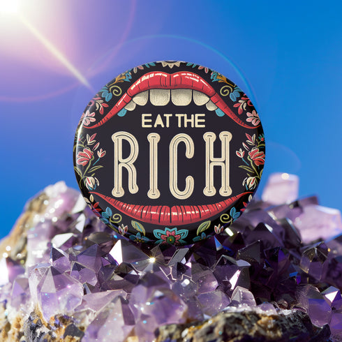 A round pin-back button with a bold, embroidered-style design featuring the phrase "EAT THE RICH" in large, vintage-style lettering. The typography is framed by a set of open red lips with detailed stitching, revealing white teeth. Surrounding the mouth are intricate floral patterns in shades of blue, red, and green, adding a decorative, folk-art-inspired touch. The button has a black background, making the details and lettering stand out