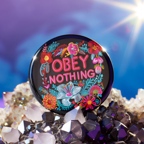 A black round pinback button featuring floral embroidery-style designs and bold red text that reads "Obey Nothing."