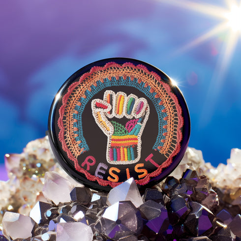 Round pinback metal button with an embroidered-style design on a black background. The center features a raised fist symbol stitched in a multicolored pattern, incorporating rainbow hues and folk-inspired textures. The fist is encircled by a vibrant, lace-like border in shades of blue, orange, and red, resembling intricate embroidery. Below the fist, the word "RESIST" is spelled out in bold, stitched-textured letters in red and orange. The button has a silver metal back with a secure pin closure.